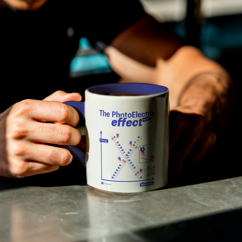 Person holding a white mug with electric blue inside and 'The Photoelectric Effect' design on a table.